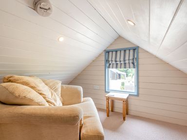 A small room with slanted white wooden walls a beige couch with cushions and a small wooden table by a window with striped blinds at The Nook in Louth