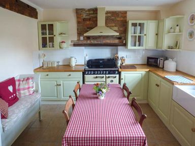A kitchen with a wooden table covered with a red and white checkered tablecloth surrounded by wooden chairs a couch with cushions green cabinets a stove and a sink at Woodside Cottage in Brompton-by-Sawdon