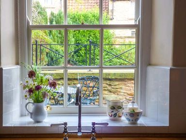A kitchen window with a sink and faucet with a white pitcher holding flowers and ceramic containers on the windowsill with a view of an outdoor patio with metal chairs and greenery at Woodside Cottage in Brompton-by-Sawdon