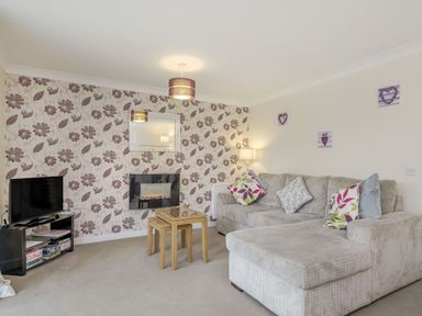 A living room with a beige sectional sofa, patterned wallpaper, a wall-mounted mirror, a fireplace, a TV on a stand, nested wooden tables, and wall art at Kymin View Monmouth