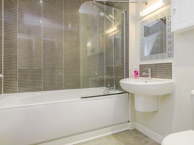 A bathroom with a bathtub and shower area with brown tiled walls a white sink with a mirror above and a soap dispenser at Kymin View in Monmouth