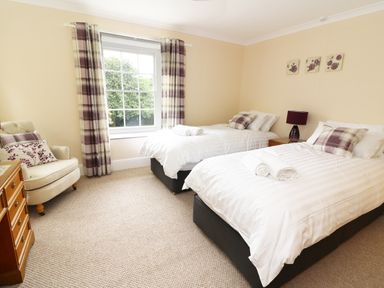 A bedroom with two single beds, a window with plaid curtains, an armchair, and a wooden dresser at Dolau Farmhouse in Lampeter
