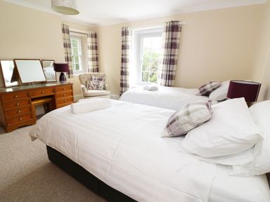A bedroom with two beds a wooden dressing table an armchair and windows with plaid curtains at Dolau Farmhouse in Lampeter
