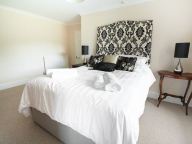 A bedroom with a bed with patterned headboard two bedside tables with lamps and rolled towels on the bed at Dolau Farmhouse in Lampeter