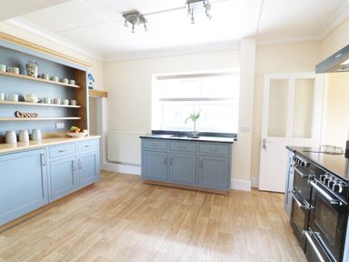 A kitchen with blue cabinets wooden shelves cups a fruit bowl a window with flowers and a black oven at Dolau Farmhouse in Lampeter