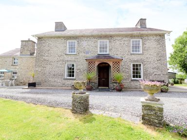 Exterior view of a stone farmhouse with a wooden porch and gravel driveway at Dolau Farmhouse in Lampeter