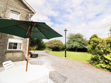 An outdoor patio area with a white table and chairs under a large green umbrella next to a stone house and a gravel driveway with grass and trees at Dolau Farmhouse in Lampeter
