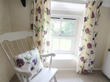 A white wooden chair with a floral cushion next to a window with floral curtains at Dolau Farmhouse in Lampeter