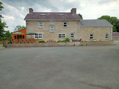 A stone farmhouse with multiple windows and outdoor seating areas including tables with umbrellas at Dolau Farmhouse in Lampeter