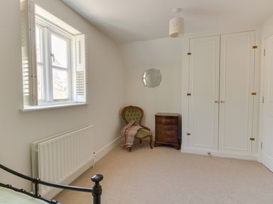 A bedroom with a window, radiator, green upholstered chair with a blanket, wooden chest of drawers, built-in wardrobe, and a decorative wall mirror at The Old School House in Piddletrenthide