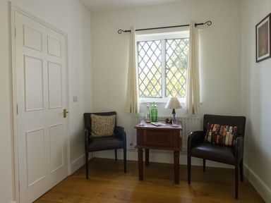 A small room with two chairs a wooden side table a window with curtains and a door at The Old School House in Piddletrenthide