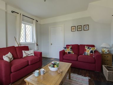 A living room with two red sofas a wooden coffee table with mugs and a fruit bowl a window with curtains and a wooden side table with a lamp at The Old School House in Piddletrenthide