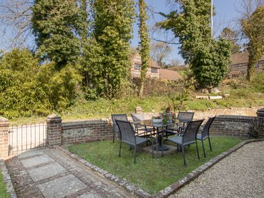 An outdoor seating area with a glass table and six chairs on grass next to a brick wall and pathway at The Old School House in Piddletrenthide