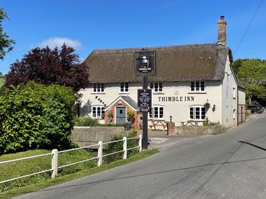 A building with a thatched roof and sign reading The Thimble Inn beside a road with bushes at The Old School House in Piddletrenthide
