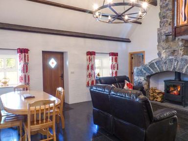 A living room with a wooden dining table and chairs a stone fireplace with firewood and a wood stove black leather sofas and floral curtains at The Well House in Kinvara County Galway