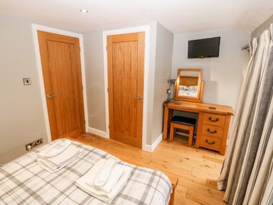 A bedroom with a bed, two wooden doors, a wooden dressing table with a mirror, a stool, a lamp, a mounted television, and striped curtains at Haddef in Nefyn