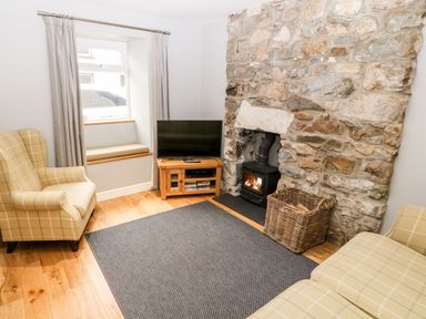 A sitting room with a TV on a wooden stand a stone fireplace with a wood burner and two checked armchairs at Haddef in Nefyn