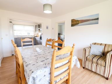 A dining room with a wooden table and chairs a striped armchair and a kitchen in the background at Sealyons Fairbourne near Dolgellau