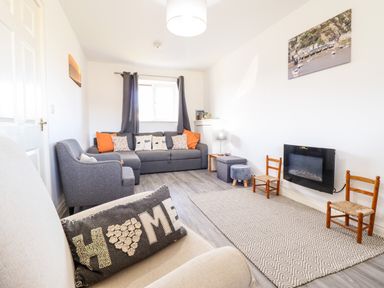 A living room with a gray sofa and armchair two wooden chairs a wall-mounted fireplace and a window with curtains at Sealyons Fairbourne near Dolgellau