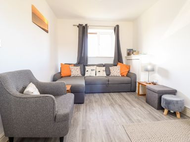 A living room with a grey armchair a grey sectional sofa with cushions and orange pillows grey curtains a small wooden table with a lamp and books a small grey ottoman and a patterned rug at Sealyons Fairbourne near Dolgellau