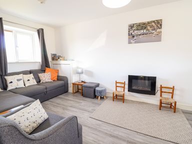 A living room with a gray sofa and armchair pillows a small table and chairs a wall mounted fireplace and a rug at Sealyons in Fairbourne near Dolgellau