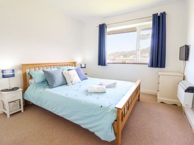 A bedroom with a wooden double bed blue bedspread and pillows bedside tables with lamps window with blue curtains and a wall-mounted TV at Sealyons in Fairbourne near Dolgellau