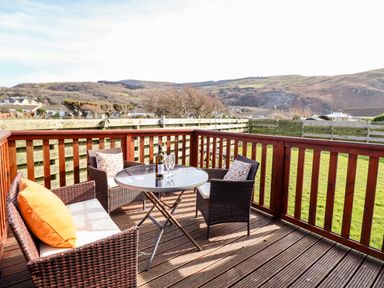 An outdoor wooden deck with wicker furniture and a bottle with glasses on a round table at Sealyons in Fairbourne near Dolgellau