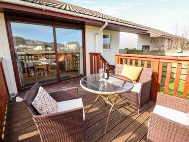 A wooden outdoor deck with a round glass table and wicker chairs with cushions at Sealyons Fairbourne near Dolgellau