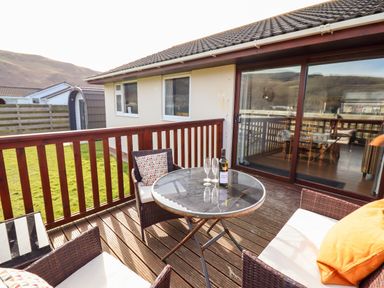 A wooden deck patio with a round glass table and wicker chairs outside a house at Sealyons Fairbourne near Dolgellau