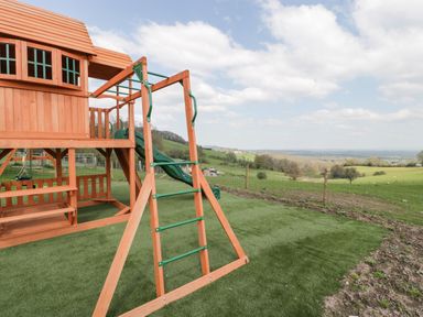 A wooden play structure with a slide and climbing ladder in a grassy yard overlooking fields at Moors Farm in Littledean