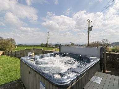 An outdoor hot tub with two glasses and a bottle on the edge overlooking a grassy field and a rural landscape at Moors Farm in Littledean