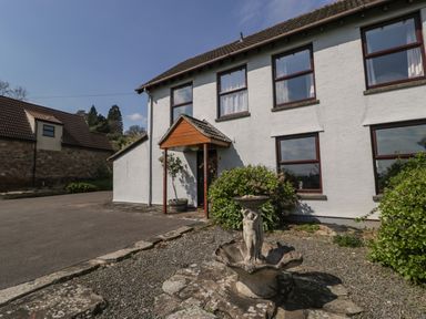 A white two-story house with brown framed windows and a small porch next to a stone fountain and bushes at Moors Farm in Littledean