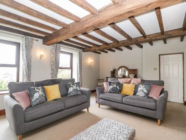 A living room with two gray sofas with cushions a patterned ottoman wooden beams on the ceiling and light curtains on windows at Moors Farm in Littledean
