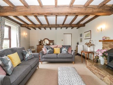 A living room with two grey sofas, patterned cushions, wooden beams on the ceiling, a stone fireplace with flowers, and a wooden sideboard at Moors Farm in Littledean