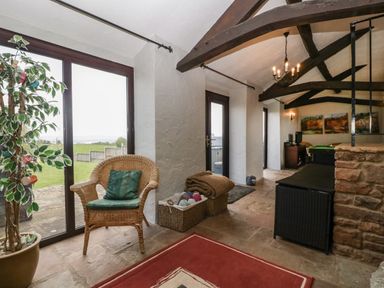 A room with wicker chair and cushion near large windows and doors overlooking a garden at Moors Farm in Littledean