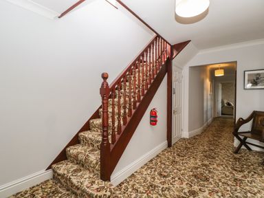 A hallway with carpeted stairs and patterned carpet on the floor and wall with a fire extinguisher and wooden chair at Moors Farm in Littledean