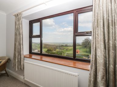 A window with curtains overlooking green fields and a red playhouse at Moors Farm in Littledean