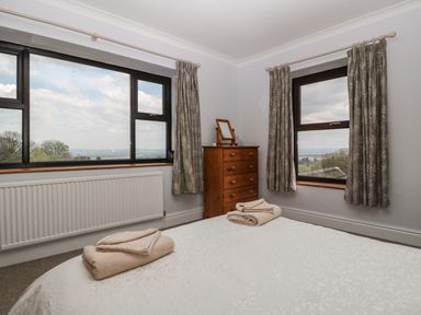 A bedroom with a bed, two folded towels on the bed, a wooden chest of drawers with a mirror, two windows with curtains at Moors Farm in Littledean