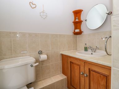 A bathroom with a toilet, a wooden cabinet with a sink, a round mirror, and small wall shelves at Moors Farm in Littledean
