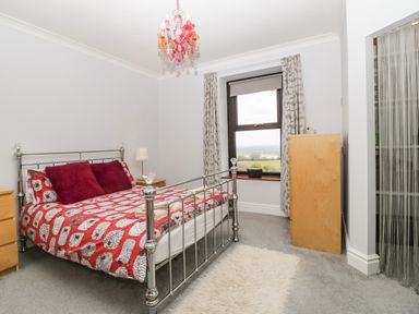 A bedroom with a metal bed frame a red patterned bedspread two red pillows two wooden dressers a window with curtains and a chandelier at Moors Farm in Littledean