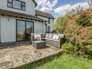 An outdoor patio with a wicker sectional sofa and table with glasses and a bottle next to a white house at Moors Farm in Littledean