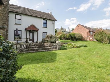 A white house with dark window frames a wooden picnic table and a large garden at Moors Farm in Littledean