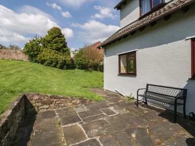 A stone patio with a bench next to a white house and a green lawn with bushes and trees at Moors Farm in Littledean