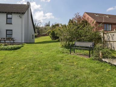A grassy backyard with a black bench near bushes and two houses in the background at Moors Farm in Littledean