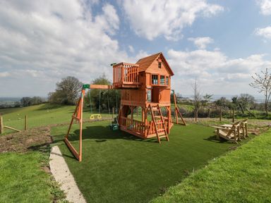 A wooden children's playhouse with swings and slides on a grassy area at Moors Farm in Littledean