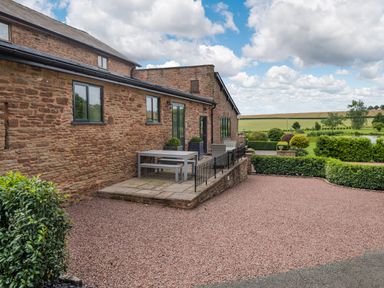 An outdoor patio area with stone tiles and a table with benches next to a brick building with a gravel driveway and manicured hedges at Teal Barn Annexe in St Weonards