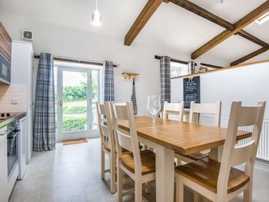 A dining area with a wooden table and six chairs next to glass doors and windows with plaid curtains at Teal Barn Annexe in St Weonards