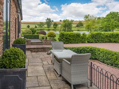 A stone patio with a glass table and wicker chairs overlooking a garden and pond at Teal Barn Annexe in St Weonards
