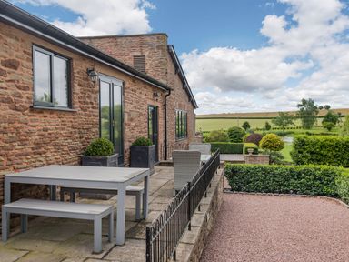 A stone patio with a gray table and benches and gray chairs overlooking a garden and fields at Teal Barn Annexe in St Weonards