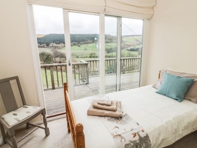A bedroom with a single bed folded towels a chair and sliding glass doors opening to a wooden deck with a view of hills at Middle Hivron in Bleddfa